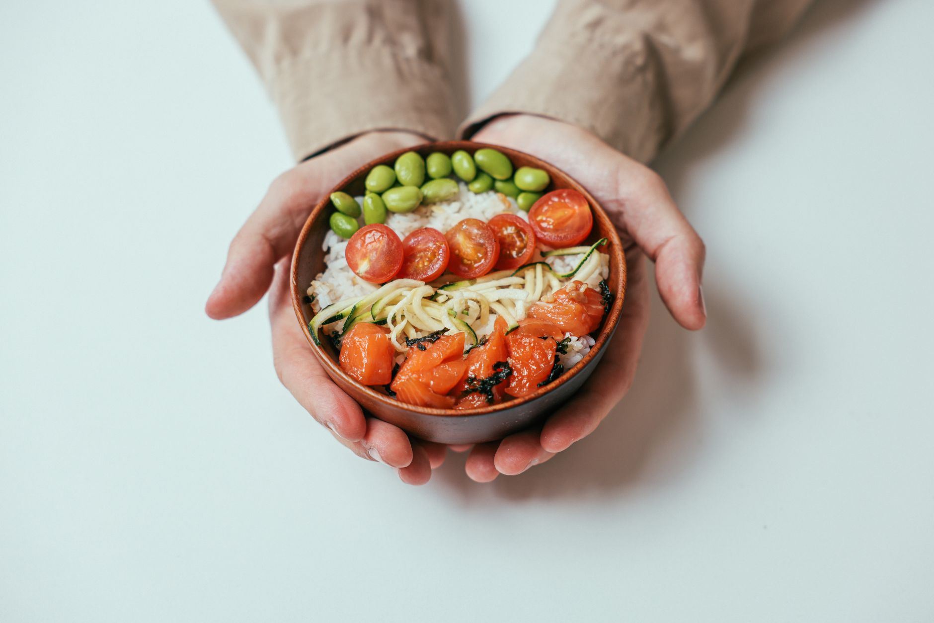 Hands holding a balanced bowl with salmon, rice, edamame and tomatoes