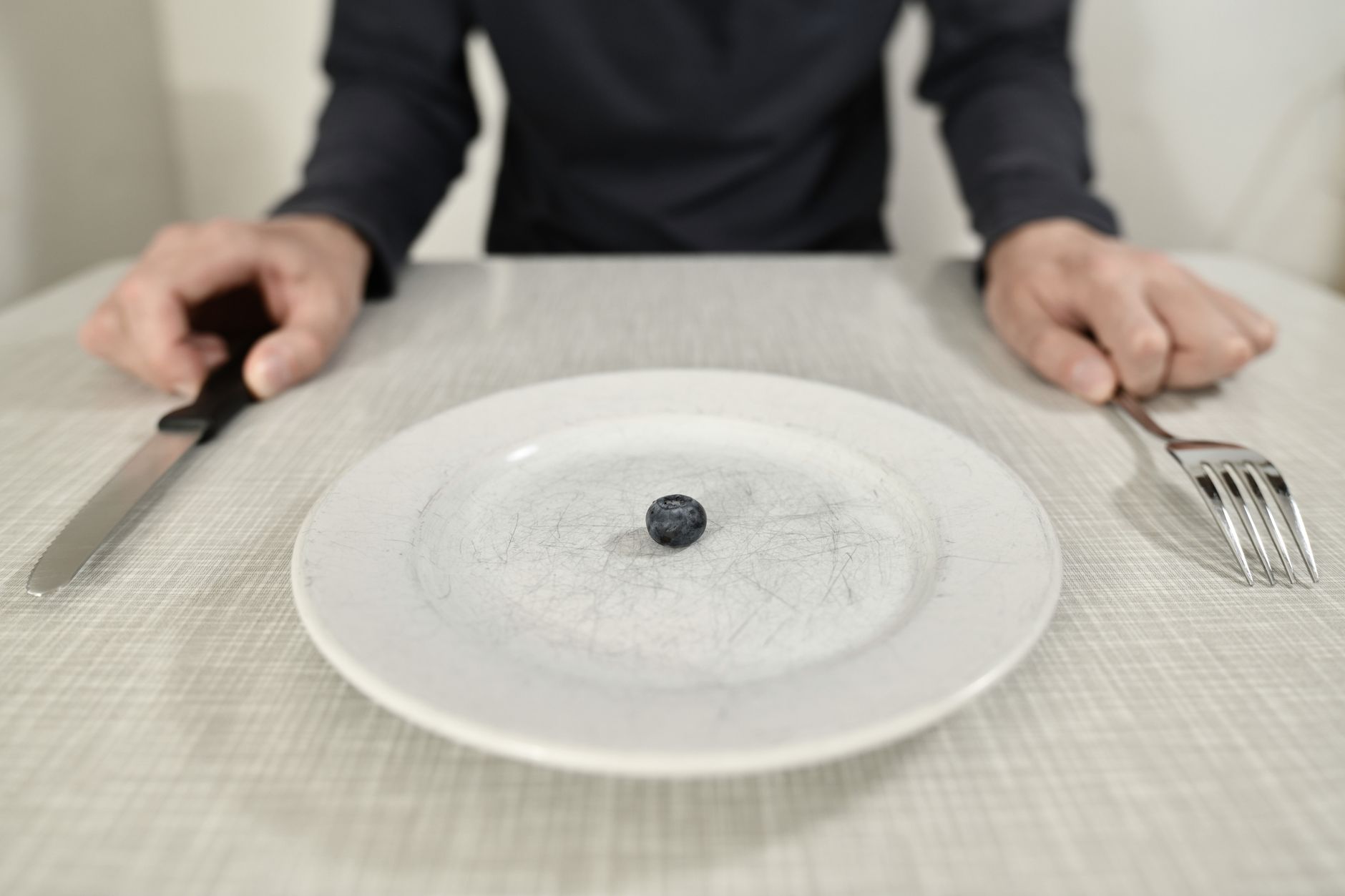 Person at a table with cutlery and a plate holding a single blueberry, illustrating the AIP elimination phase