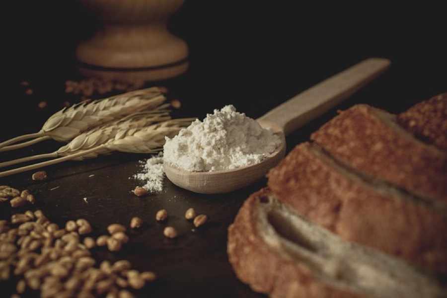 Wheat ears, loose wheat grains and a wooden spoon of white flour beside a loaf of rustic bread