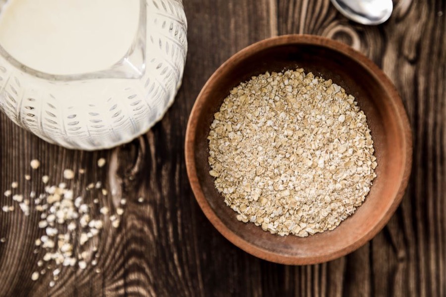 Glass jug of oat milk beside a wooden bowl of rolled oats on a dark wood table
