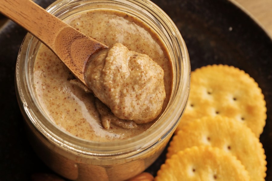 A glass jar of almond butter flecked with brown almond skins, with a wooden spoon and whole almonds