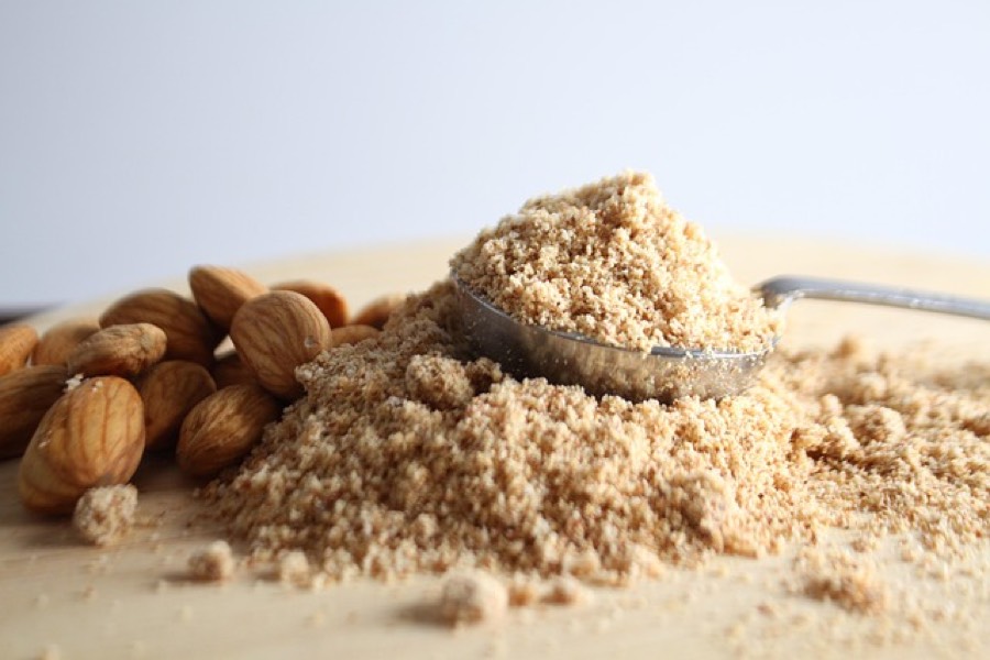 A heap of finely ground almond flour with a spoon in it and whole almonds beside it on a wooden board