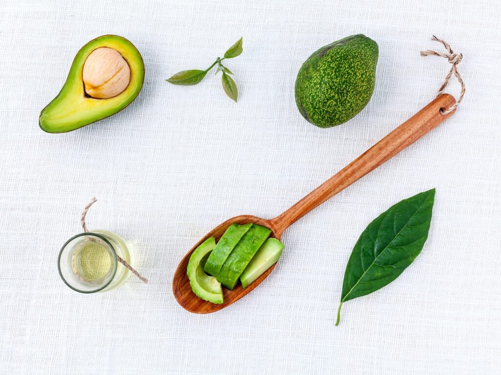 Small glass jar of avocado oil beside a halved avocado, sliced avocado on a wooden spoon and avocado leaves