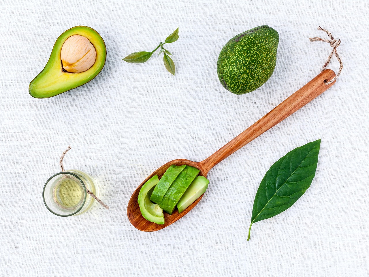 Small glass jar of avocado oil beside a halved avocado, sliced avocado on a wooden spoon and avocado leaves
