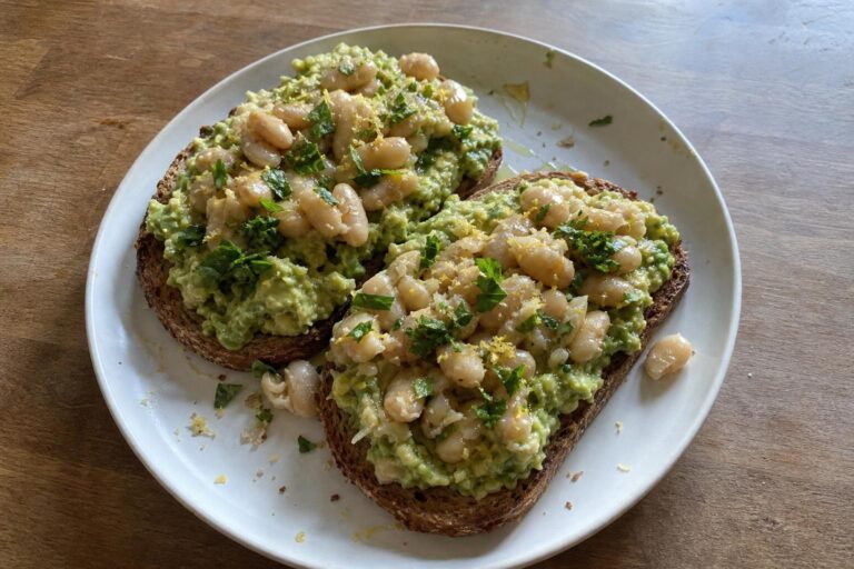 Two slices of toasted whole grain sourdough topped with lumpy mashed avocado, warm smashed cannellini beans, grated lemon zest and chopped parsley on a plate.