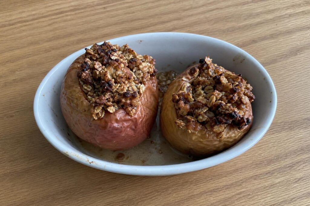 Two baked apples stuffed with a toasted walnut and oat filling in a small baking dish, skins wrinkled and juices pooling around the base.