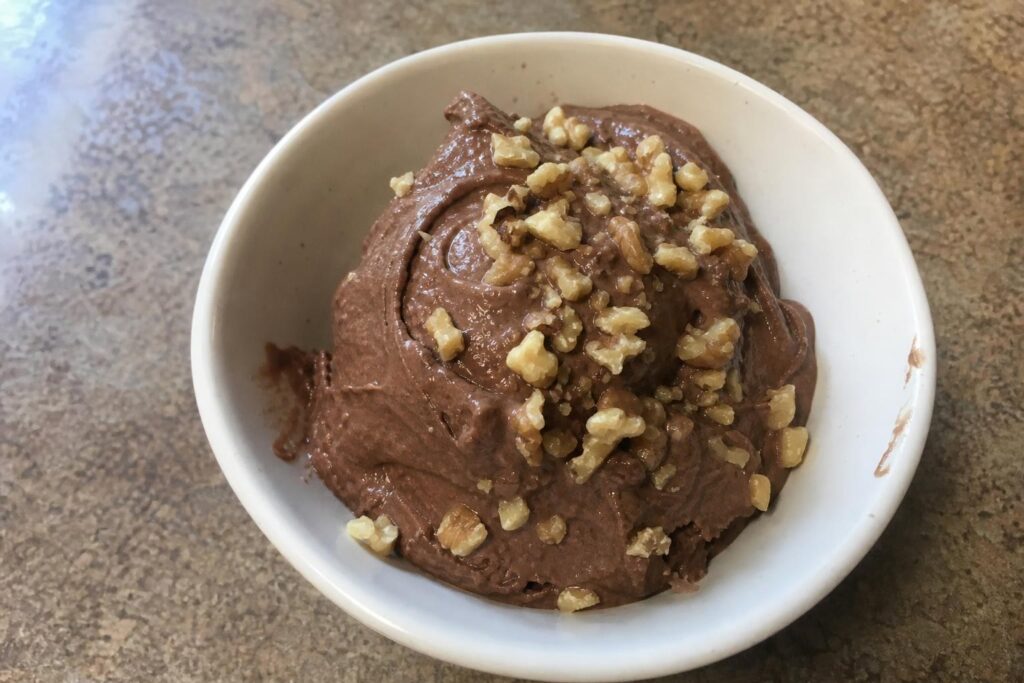 A single bowl of banana cocoa nice cream, thick dark brown chocolate soft-serve swirls scooped in an irregular mound, scattered unevenly with chopped walnut pieces, slightly melting at the edges.
