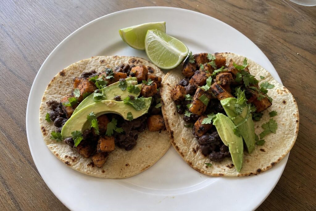 Two soft corn tortillas filled with lightly mashed black beans, roasted cubed sweet potato with caramelized edges, sliced avocado, and chopped cilantro on a white plate, with lime wedges alongside.