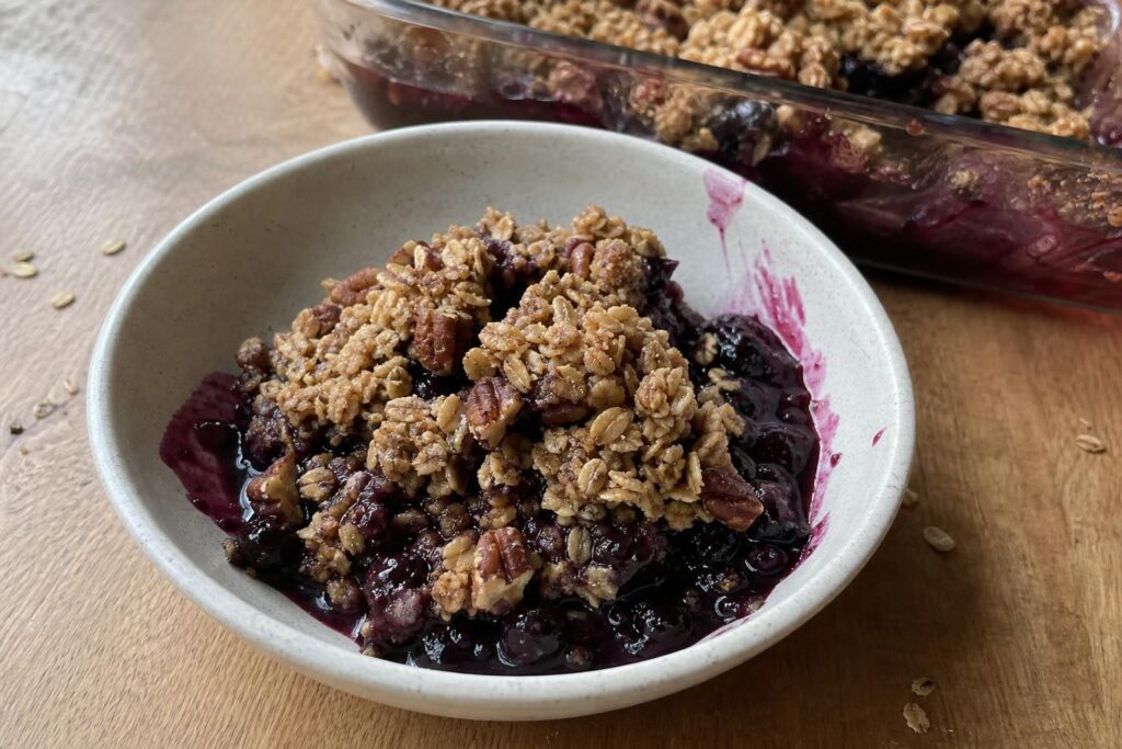 A small bowl of blueberry oat crumble with thick dark purple blueberry filling soaking through golden oat and pecan clumps, spooned out unevenly, with the glass baking dish visible in the background.