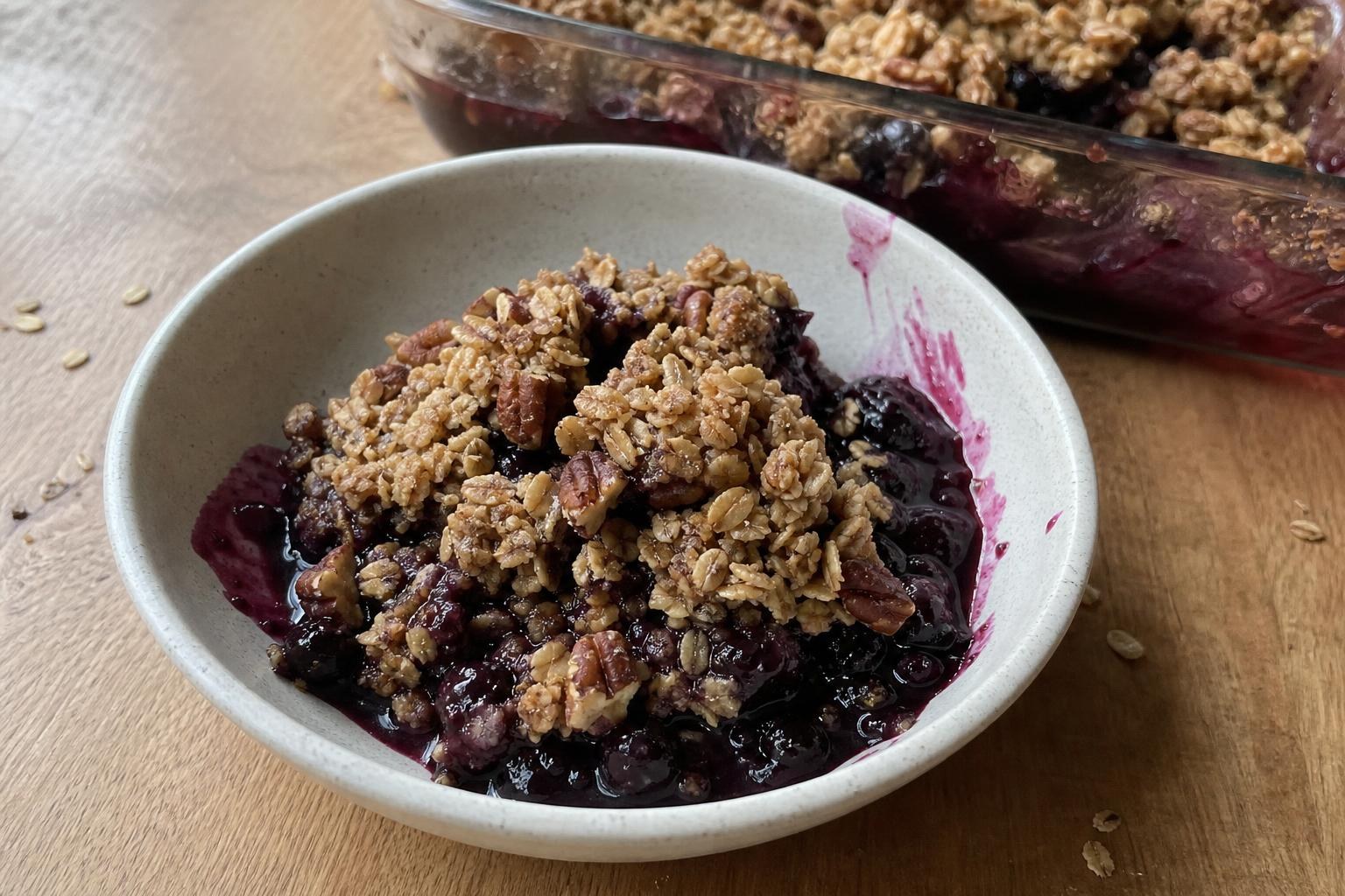 A small bowl of blueberry oat crumble with thick dark purple blueberry filling soaking through golden oat and pecan clumps, spooned out unevenly, with the glass baking dish visible in the background.