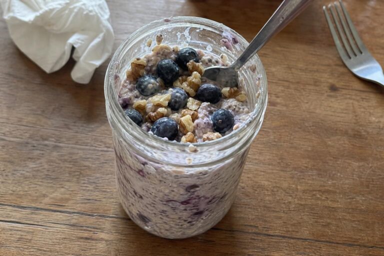 A glass jar of overnight oats with a creamy purple-flecked base, topped with a loose handful of fresh blueberries and roughly chopped walnuts, a spoon resting in the jar.