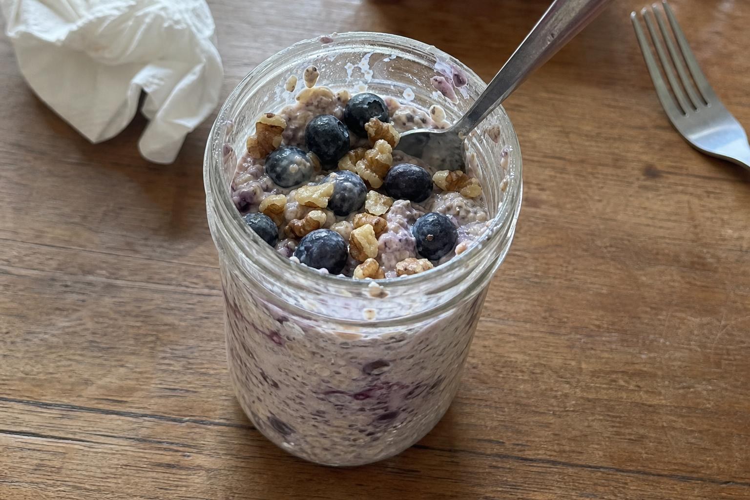 A glass jar of overnight oats with a creamy purple-flecked base, topped with a loose handful of fresh blueberries and roughly chopped walnuts, a spoon resting in the jar.