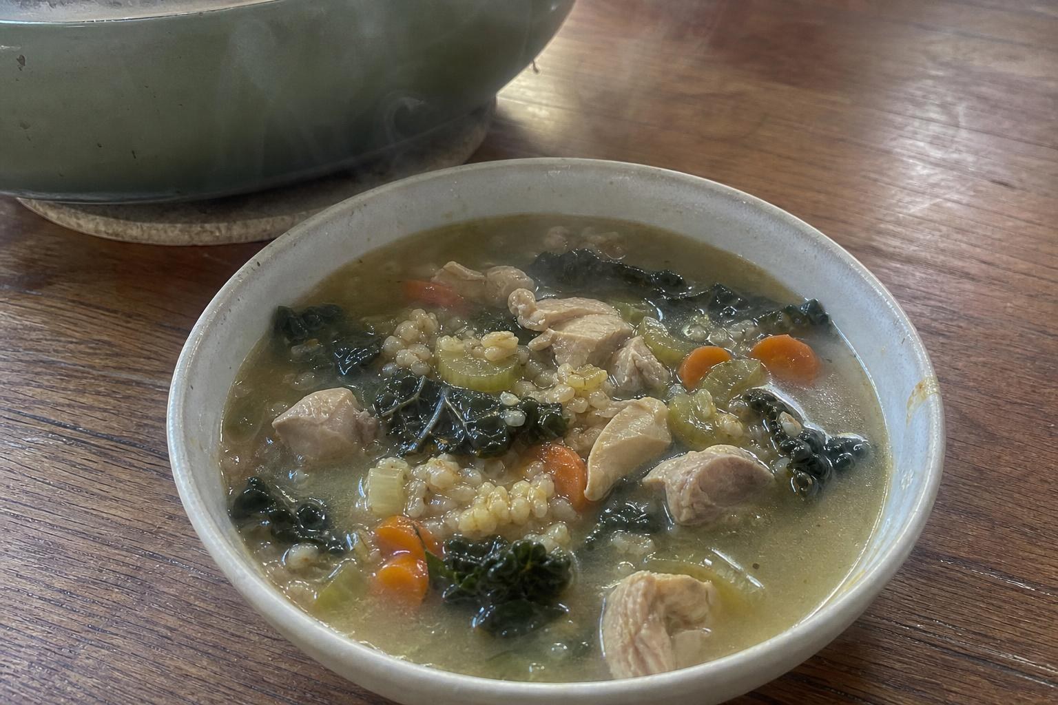 Un plato de sopa de pollo, arroz integral y col rizada en el que se ven trozos irregulares de pollo dorado, medias lunas de zanahoria, rodajas de apio, granos de arroz integral y hojas de col rizada de color verde oscuro troceadas a mano en un caldo dorado y turbio, con la olla detrás, sobre una mesa de madera.