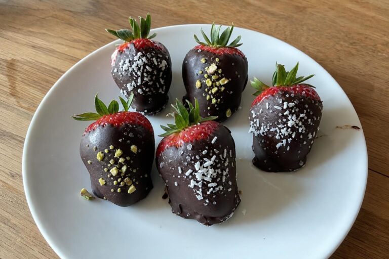 Strawberries with green stems attached, coated about three quarters of the way up in matte dark chocolate and topped with shredded coconut, chopped pistachios, and flaky sea salt, on a plain white plate.