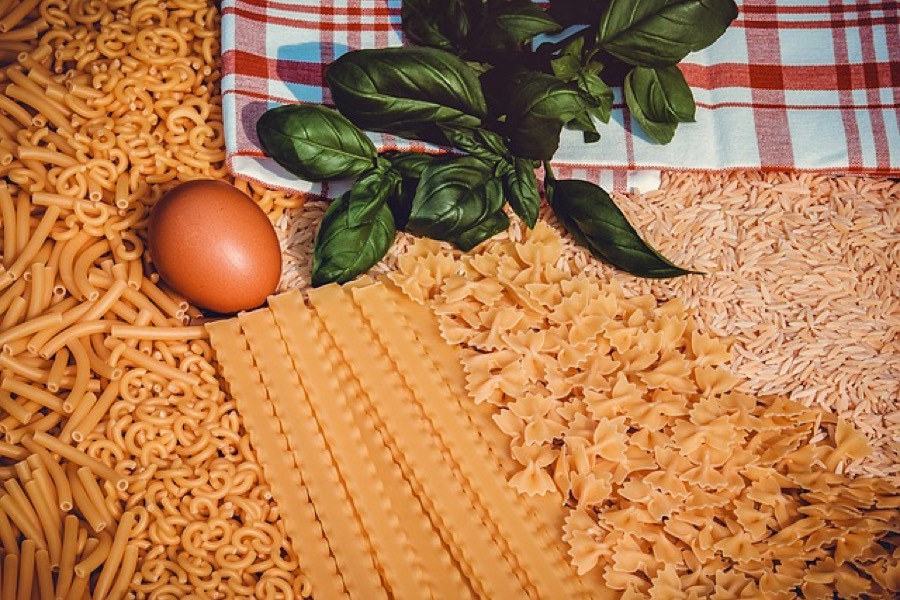 Dry pasta in several shapes, macaroni, lasagne, farfalle and orzo, laid out beside fresh basil on a checked cloth
