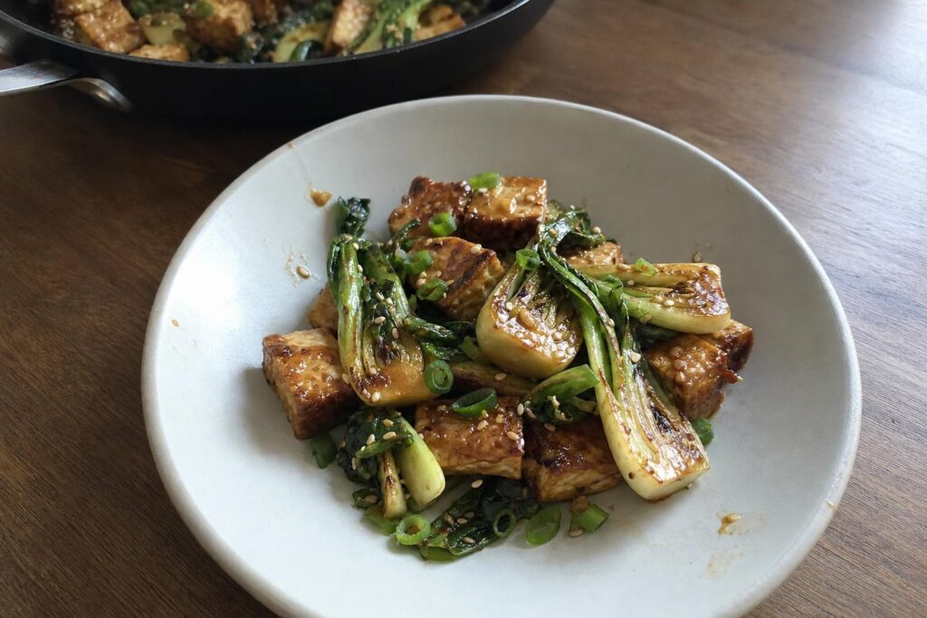 A serving of ginger sesame tofu and bok choy in a shallow bowl, showing golden-seared tofu cubes, charred baby bok choy halves, a glossy dark sauce, sesame seeds, and sliced scallions on top, with the skillet visible behind it.