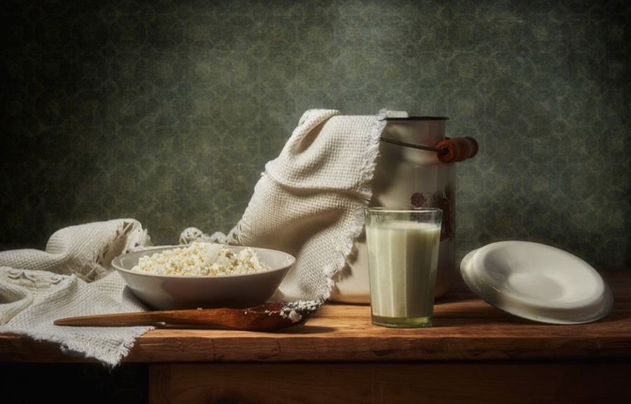 Glass of plain kefir beside a bowl of fresh curds and a milk can on a wooden table