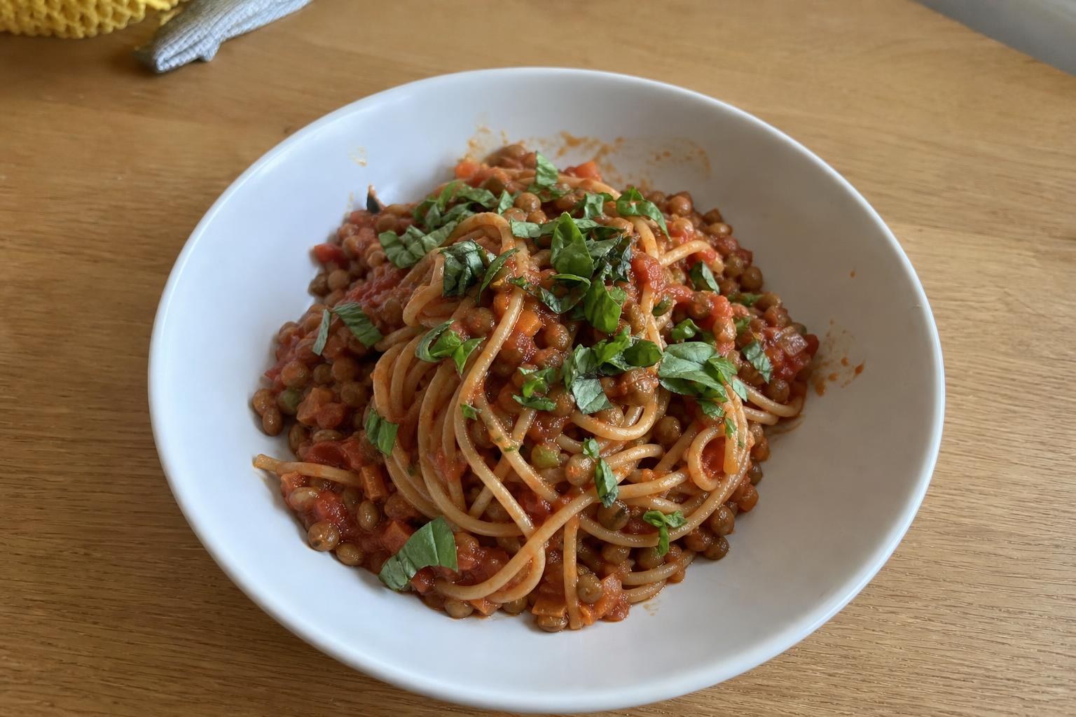 A shallow bowl of lentil bolognese tossed with whole-wheat spaghetti, showing chunks of lentils and finely diced carrot and celery in a thick tomato sauce, topped with chopped fresh basil.