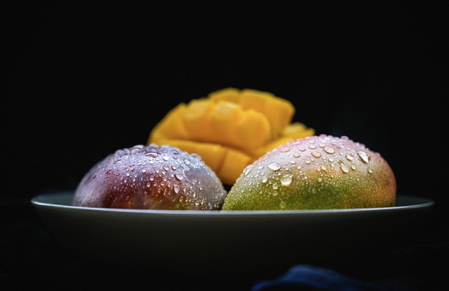 Two whole mangoes and a cut mango half showing the orange flesh, in a white bowl