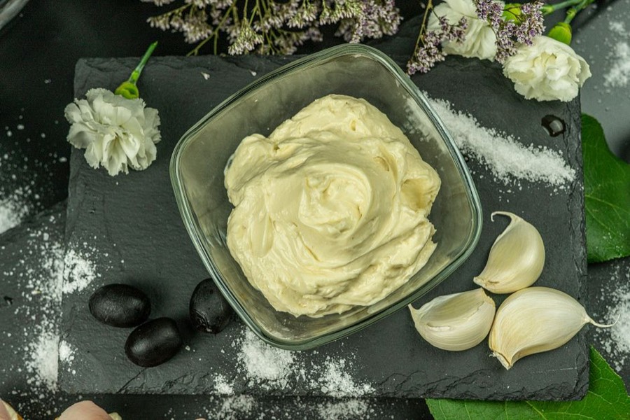 Bowl of creamy mayonnaise on a slate board next to garlic cloves and black olives