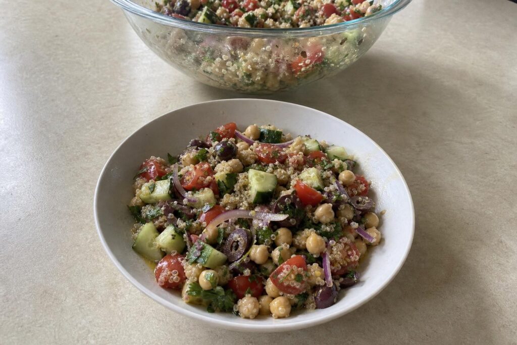 A shallow bowl of quinoa salad with chickpeas, diced cucumber, halved cherry tomatoes, thinly sliced red onion, halved Kalamata olives, and chopped parsley, tossed together and glistening with dressing.