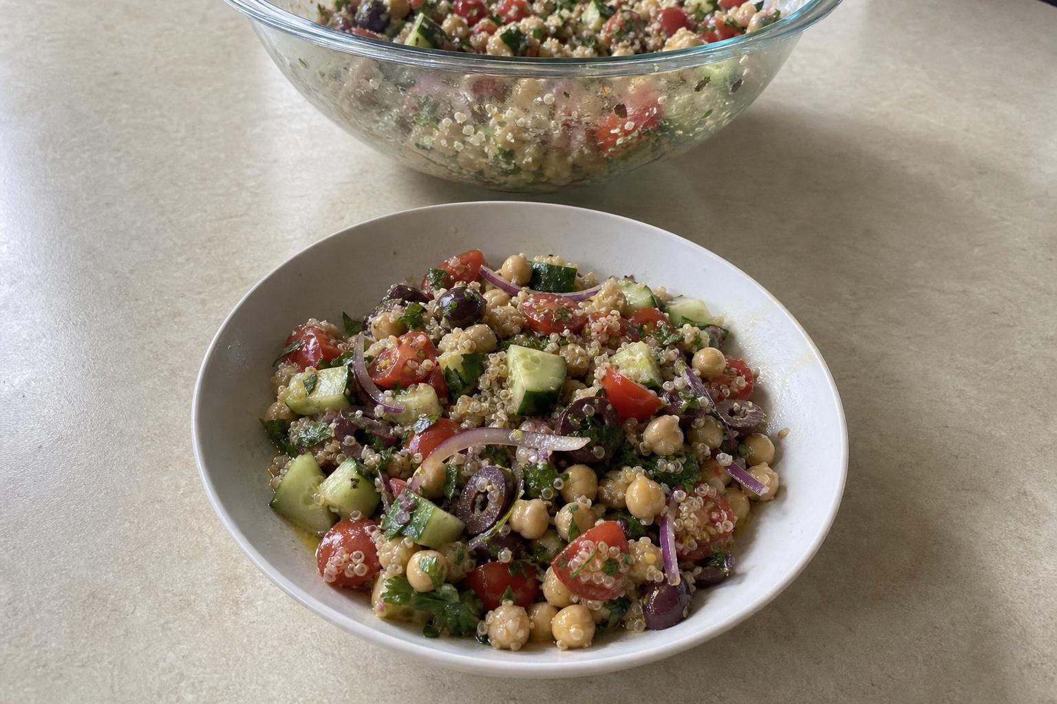 A shallow bowl of quinoa salad with chickpeas, diced cucumber, halved cherry tomatoes, thinly sliced red onion, halved Kalamata olives, and chopped parsley, tossed together and glistening with dressing.
