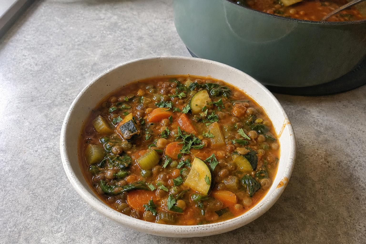 A bowl of Mediterranean lentil and vegetable soup with whole brown lentils, carrot half moons, sliced celery, zucchini quarter-rounds, and wilted spinach in a tomato-tinted broth, topped with chopped parsley.
