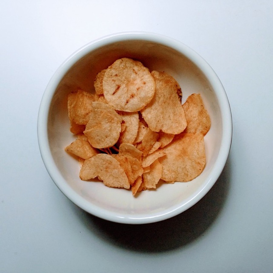 A white bowl of thin salted potato chips seen from above on a pale table