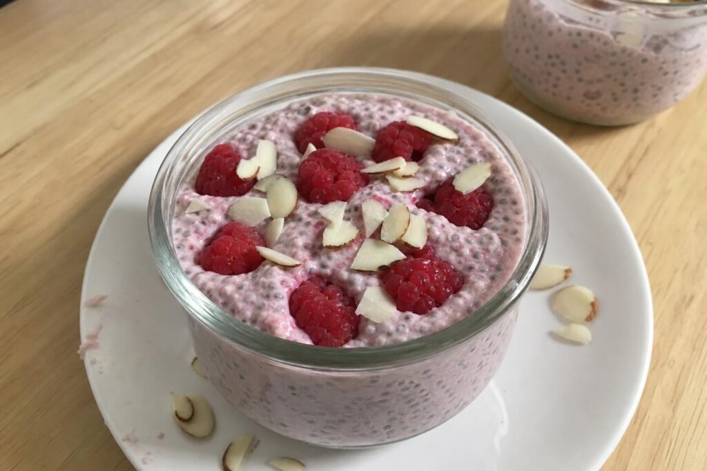 Un tarro de cristal con un espeso pudín de chía rosa salpicado de semillas de chía oscuras, colocado sobre un plato blanco y coronado con frambuesas frescas enteras y almendras crudas en láminas esparcidas de forma irregular por la superficie.