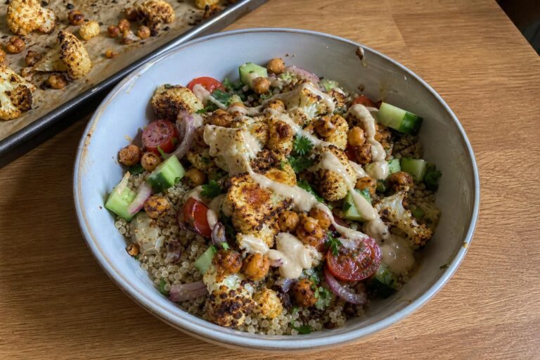 A shallow bowl of warm quinoa topped with deeply browned roasted cauliflower florets and crisp chickpeas, diced cucumber, halved cherry tomatoes, sliced red onion, a drizzle of pale tahini sauce and chopped parsley.