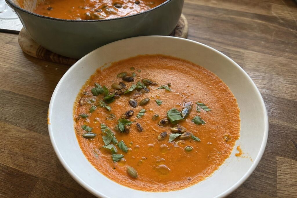 A bowl of smooth deep orange tomato and red pepper soup topped with toasted pumpkin seeds, torn fresh basil and a drizzle of olive oil.