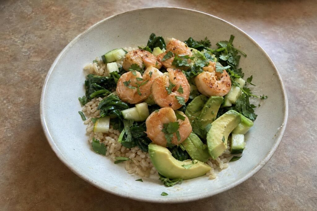 A shallow bowl of brown rice topped with wilted spinach, diced cucumber, thick avocado slices and seared pink-orange shrimp, with a light lime dressing drizzled over and chopped cilantro scattered on top.