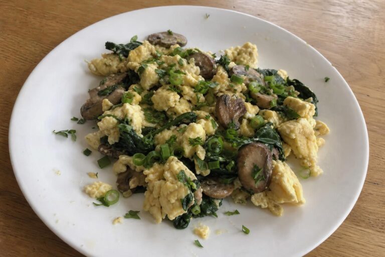 A plate of soft scrambled eggs studded with browned sliced cremini mushrooms and wilted dark green spinach, topped with chopped parsley and sliced scallion greens.
