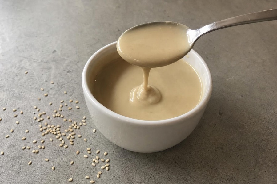 A white bowl of pale tahini with a spoon lifting a ribbon of the paste, sesame seeds scattered beside it