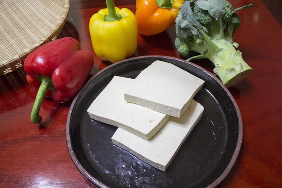 Slices of raw firm tofu on a dark plate, with red, yellow and orange peppers and a head of broccoli behind them