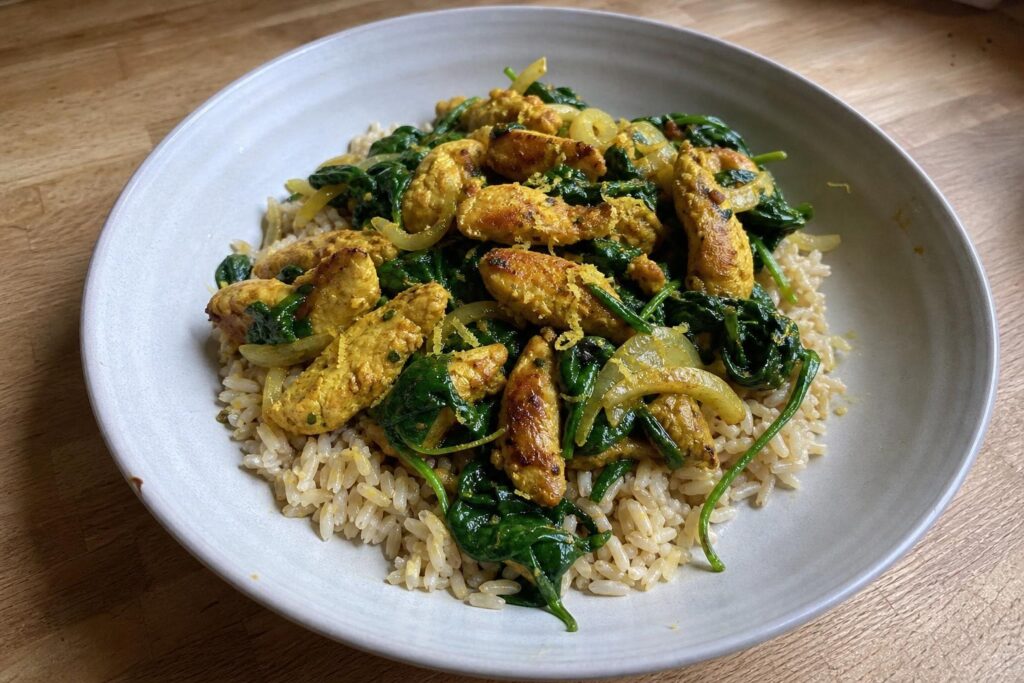 A shallow bowl of brown rice topped with golden yellow turmeric chicken strips and dark wilted spinach leaves, glossy with olive oil and lemon.