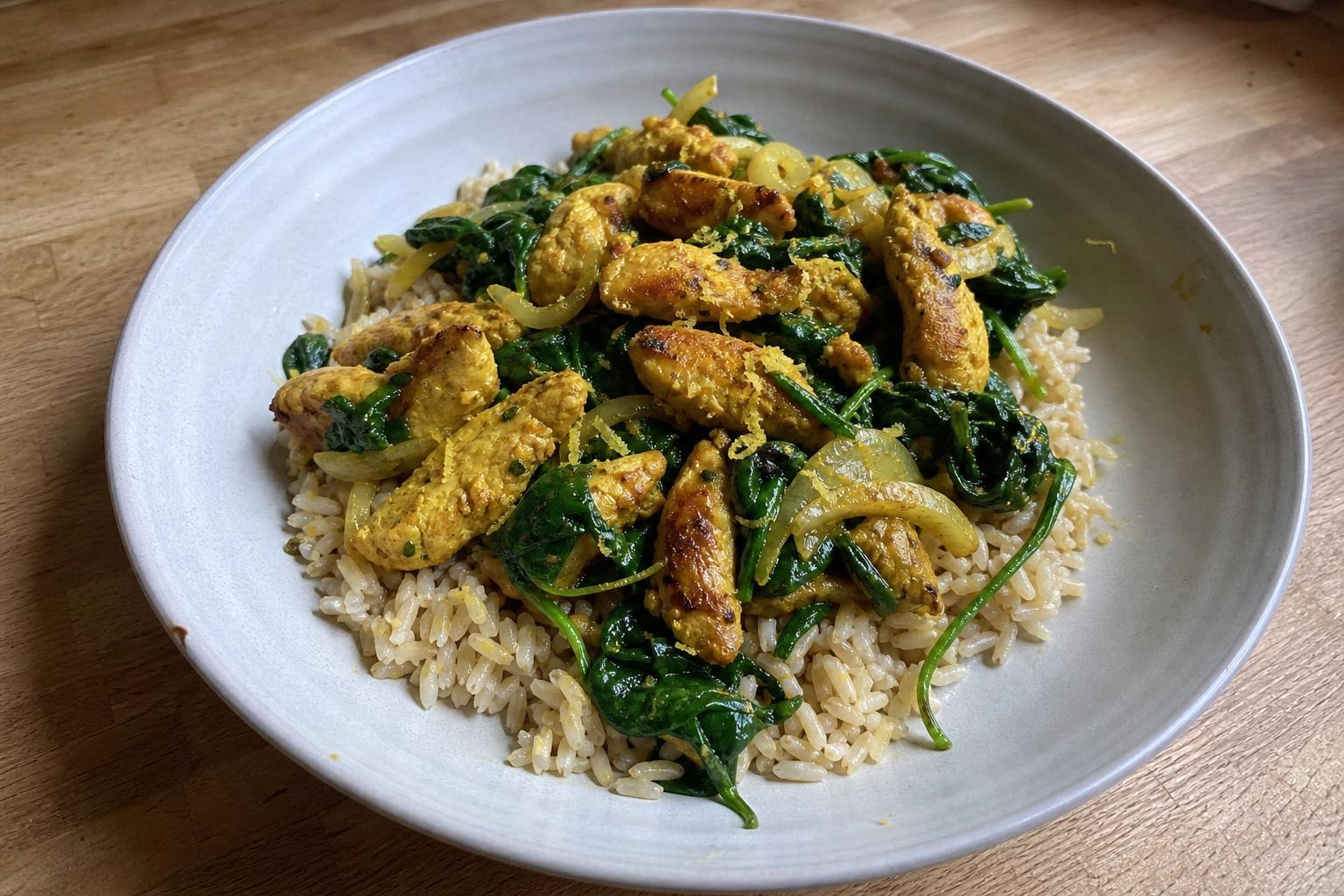 A shallow bowl of brown rice topped with golden yellow turmeric chicken strips and dark wilted spinach leaves, glossy with olive oil and lemon.
