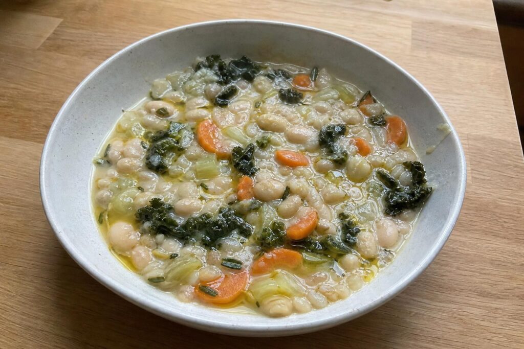 A shallow bowl of creamy white bean and rosemary stew with visible cannellini beans, carrot half-moons, celery and wilted dark kale, topped with a raw olive oil drizzle and flecks of chopped rosemary.