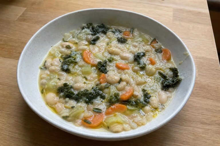 A shallow bowl of creamy white bean and rosemary stew with visible cannellini beans, carrot half-moons, celery and wilted dark kale, topped with a raw olive oil drizzle and flecks of chopped rosemary.