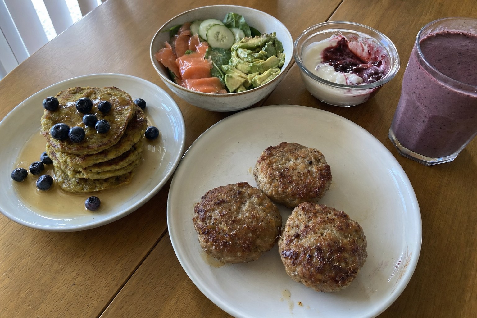 AIP breakfast ideas on a kitchen table: sausage patties, plantain pancakes with blueberries, smoked salmon bowl, coconut yogurt and a berry smoothie