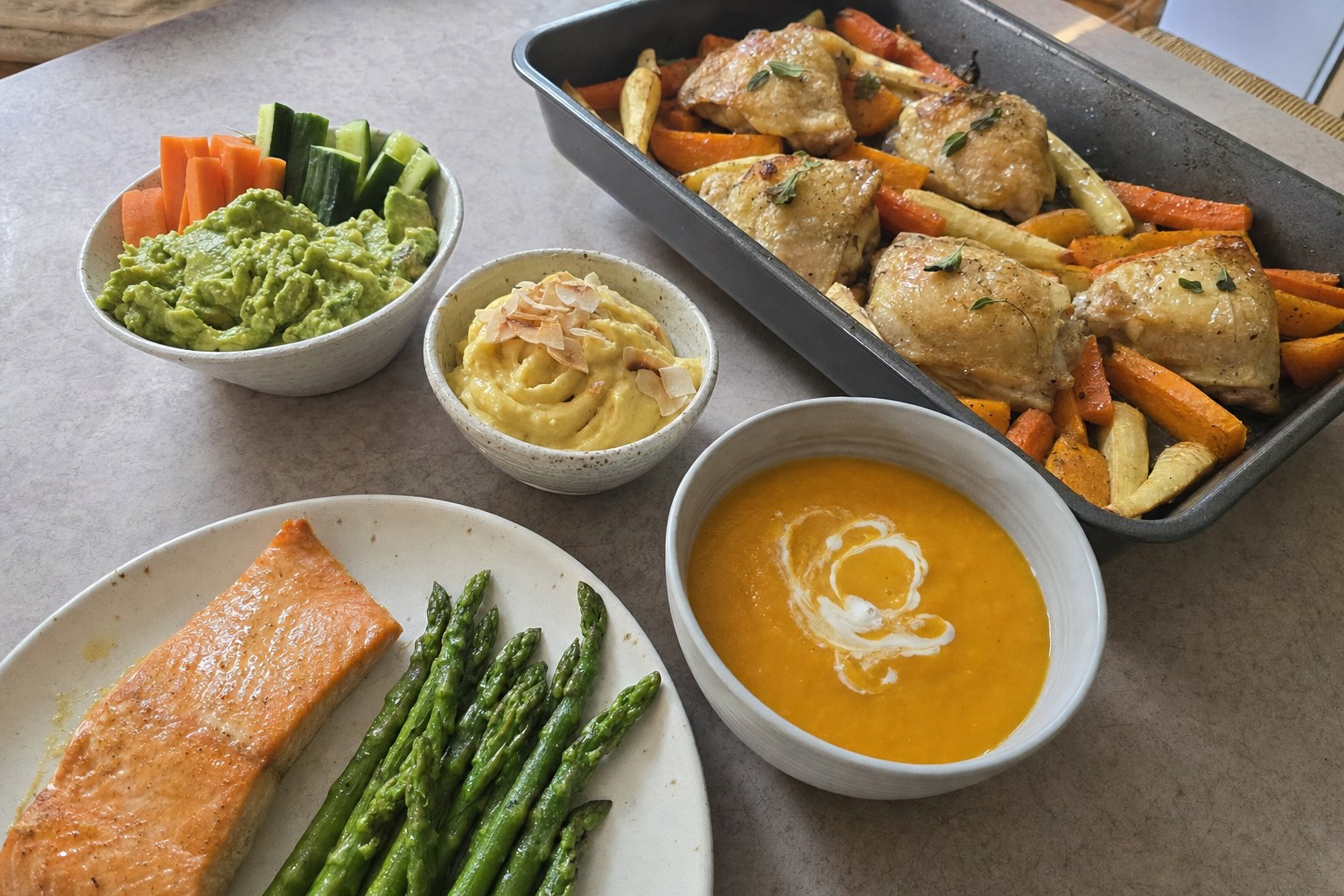 AIP recipes on a kitchen table: salmon with asparagus, roast chicken with root vegetables, carrot soup, guacamole and mango nice cream