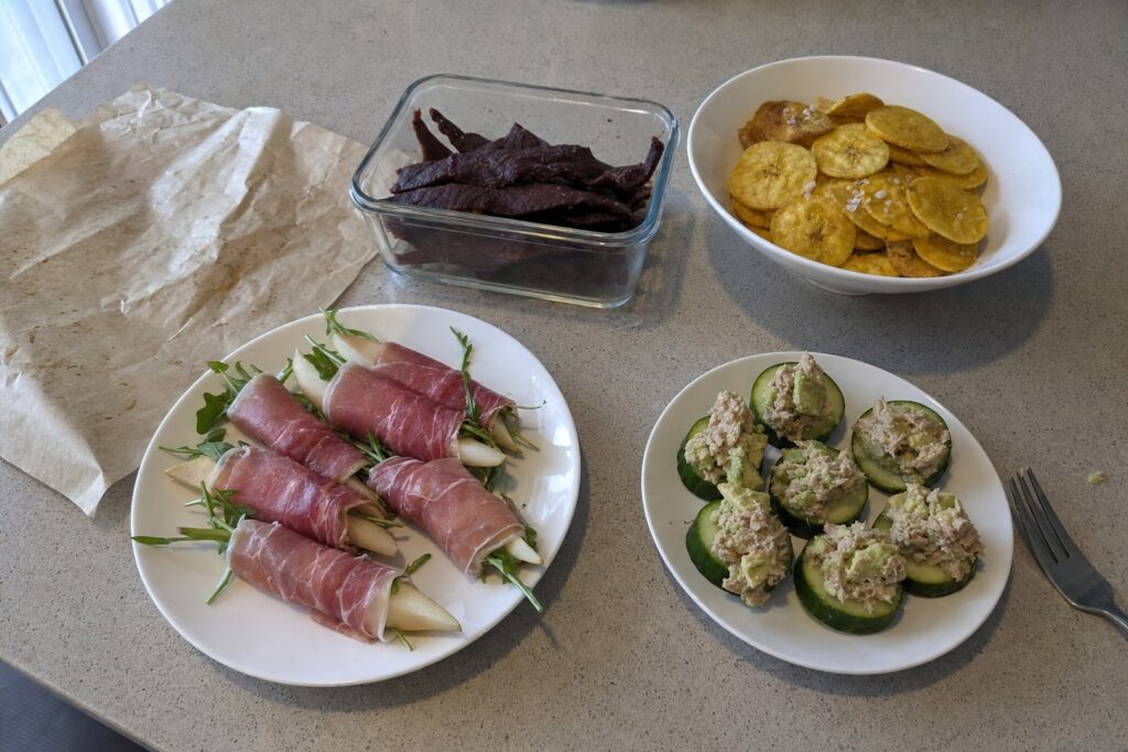 AIP snacks on a kitchen counter: homemade beef jerky, baked plantain chips, prosciutto-wrapped pears and tuna salad on cucumber rounds