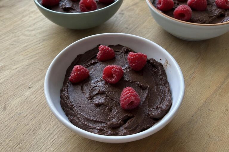 Un cuenco pequeño con mousse de chocolate y aguacate muy oscura y mate, con unas cuantas frambuesas frescas esparcidas por encima y marcas irregulares de cuchara en la superficie, sobre una mesa de cocina de las de todos los días.