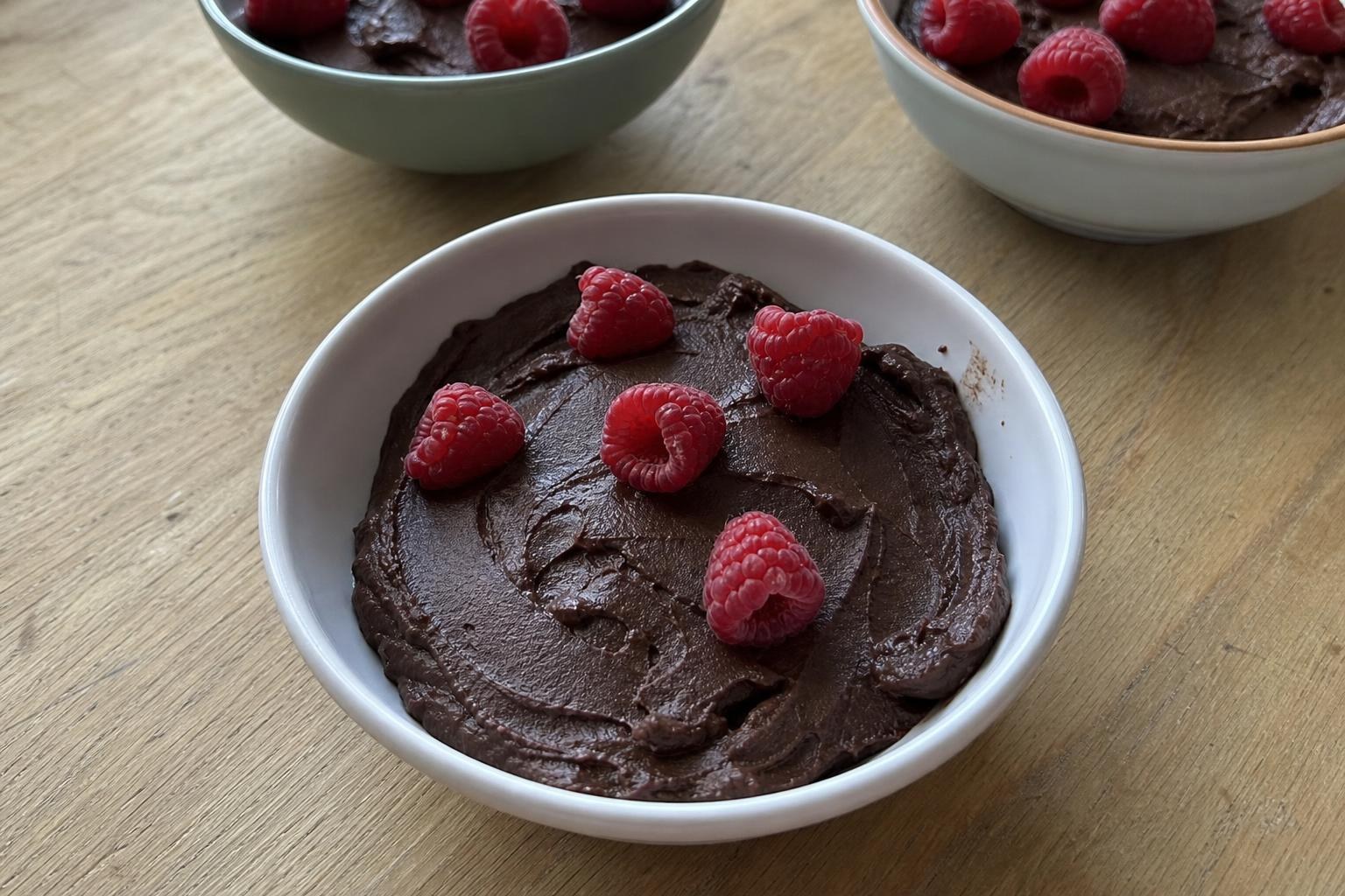 Un cuenco pequeño con mousse de chocolate y aguacate muy oscura y mate, con unas cuantas frambuesas frescas esparcidas por encima y marcas irregulares de cuchara en la superficie, sobre una mesa de cocina de las de todos los días.