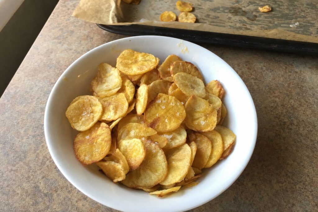 Bowl of homemade baked plantain chips with sea salt on a kitchen counter, with the baking sheet behind it