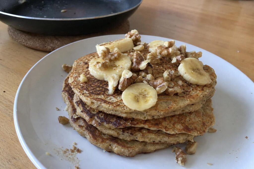 A stack of oat and banana pancakes on a white plate, topped with banana slices and chopped walnuts, with a skillet blurred in the background on a wooden table.