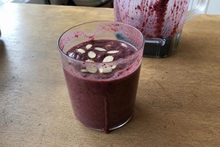 Un batido espeso de cerezas de color rojo oscuro en un vaso de los de todos los días, con almendras laminadas por encima y el vaso de la batidora difuminado al fondo.