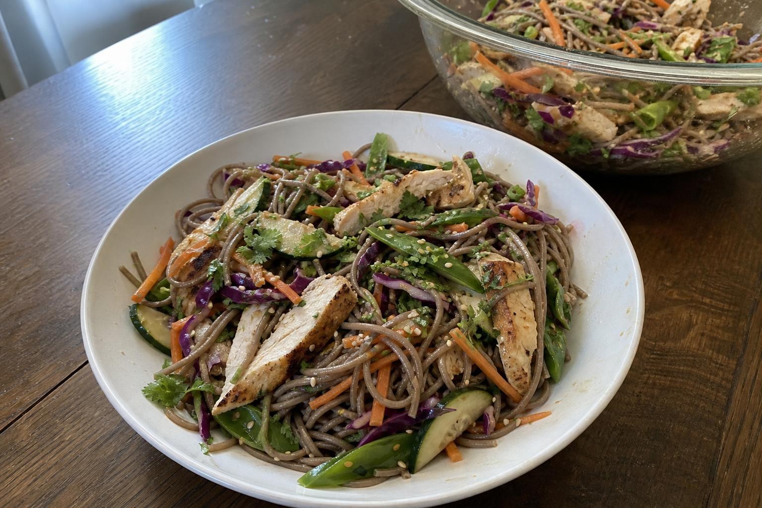A wide shallow bowl of cold soba noodle salad with seared chicken strips, matchstick carrots, red cabbage, cucumber half moons, sugar snap peas, sliced scallions, cilantro and sesame seeds.