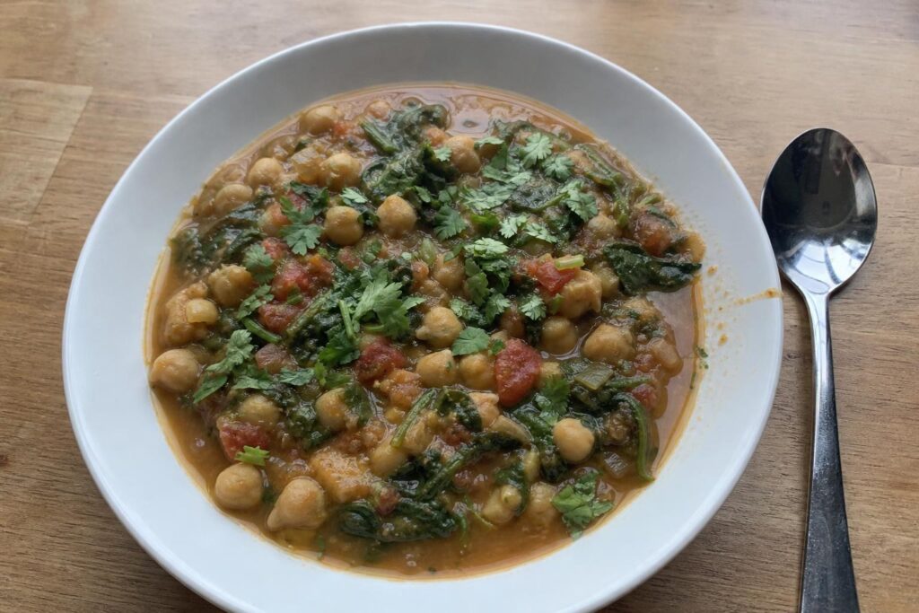 A shallow everyday bowl filled with thick chickpea and spinach stew, whole and mashed chickpeas in a cumin-colored broth, dark wilted spinach leaves, pieces of tomato and chopped cilantro scattered on top, a spoon resting beside the bowl.