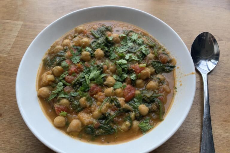 A shallow everyday bowl filled with thick chickpea and spinach stew, whole and mashed chickpeas in a cumin-colored broth, dark wilted spinach leaves, pieces of tomato and chopped cilantro scattered on top, a spoon resting beside the bowl.